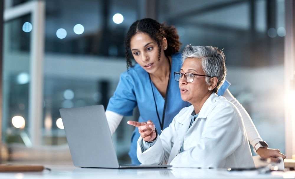 A medical professional and a nurse looking at a laptop together in a brightly lit office setting.
