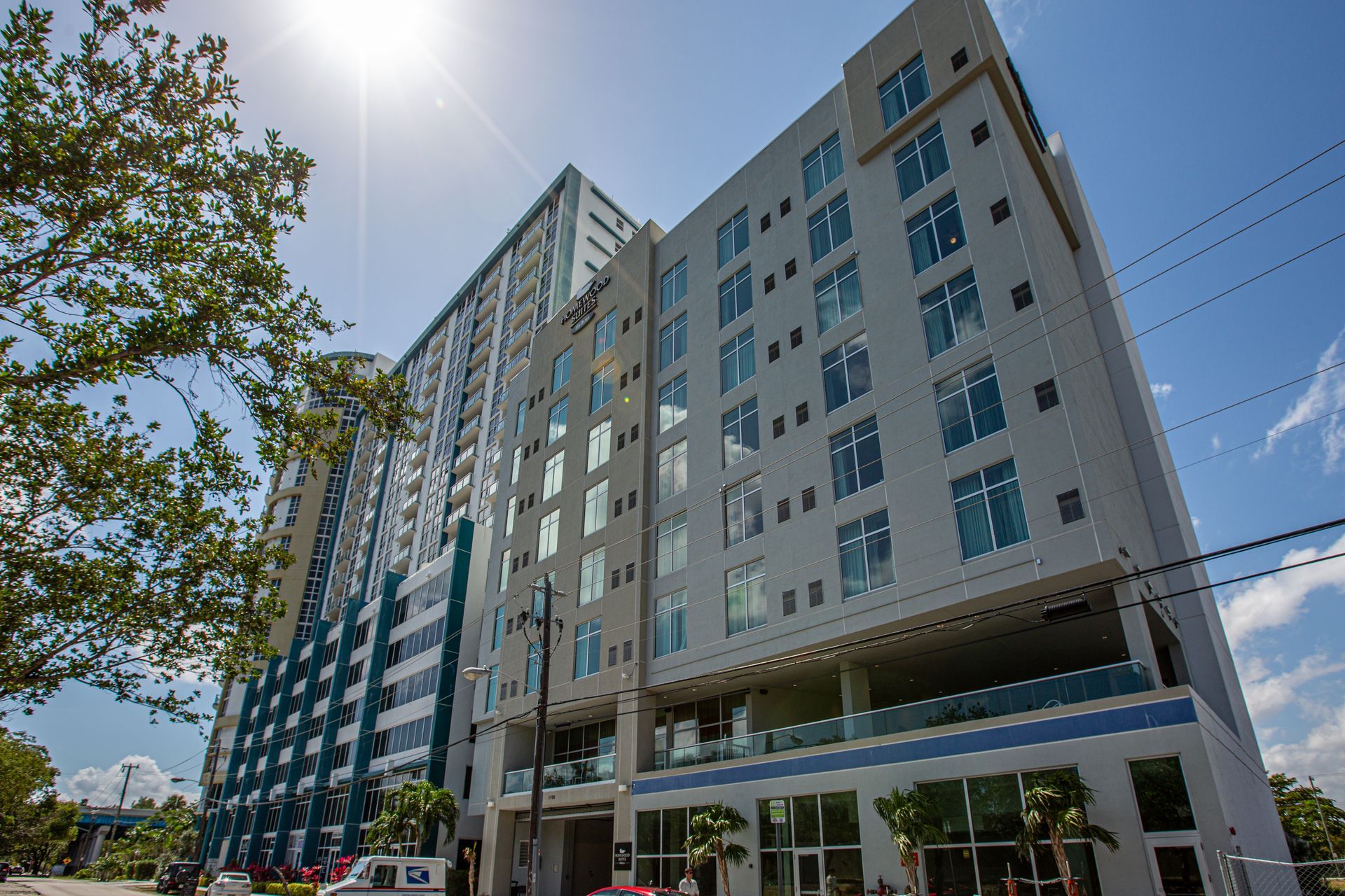 Modern high-rise building with many windows, white and blue exterior, sunny sky, power lines, and trees.