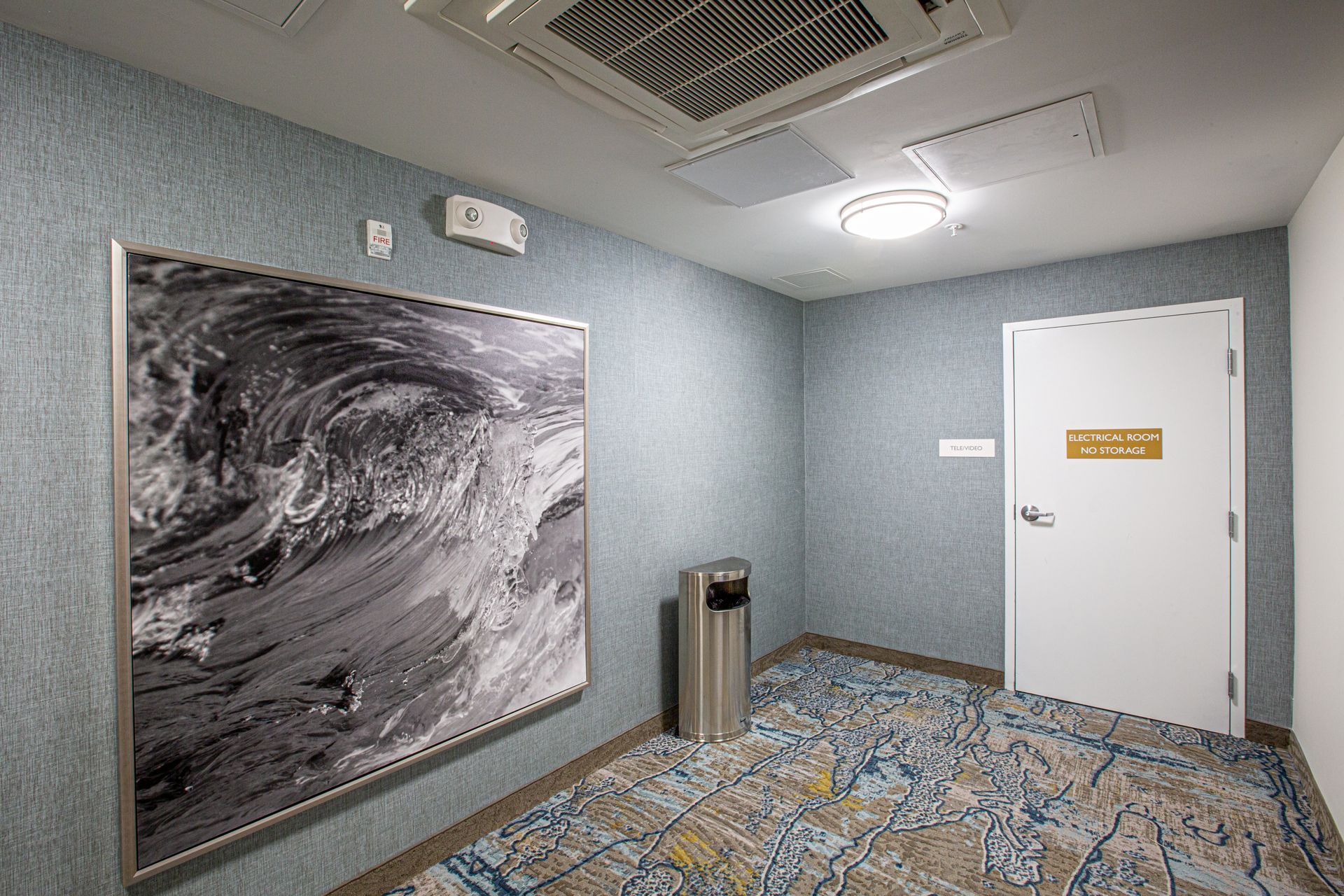 Hallway with artwork of waves, waste bin, white door, patterned carpet, and textured blue walls.