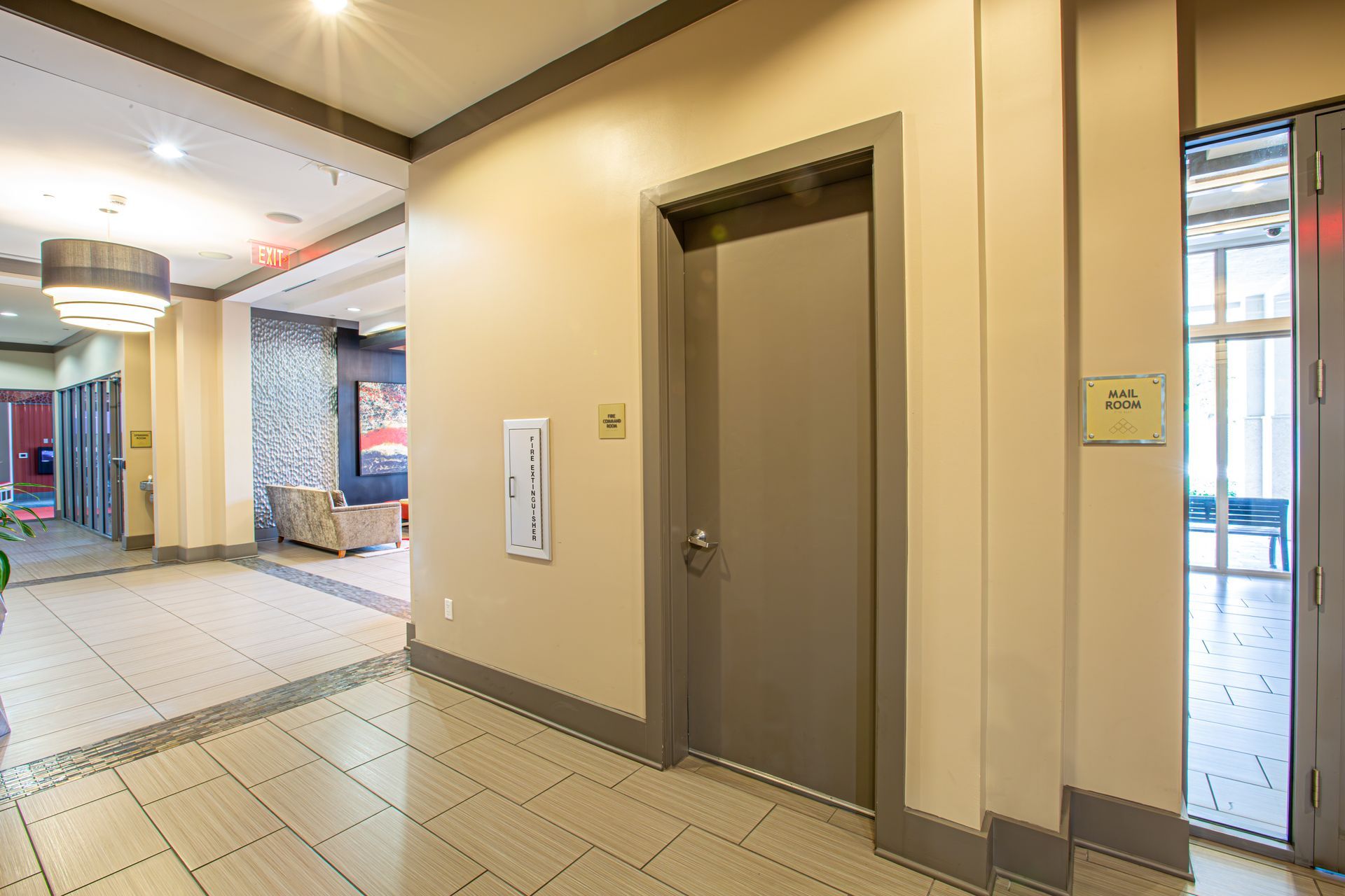 Hallway with beige walls, gray door and trim, fire extinguisher, and a doorway with windows.