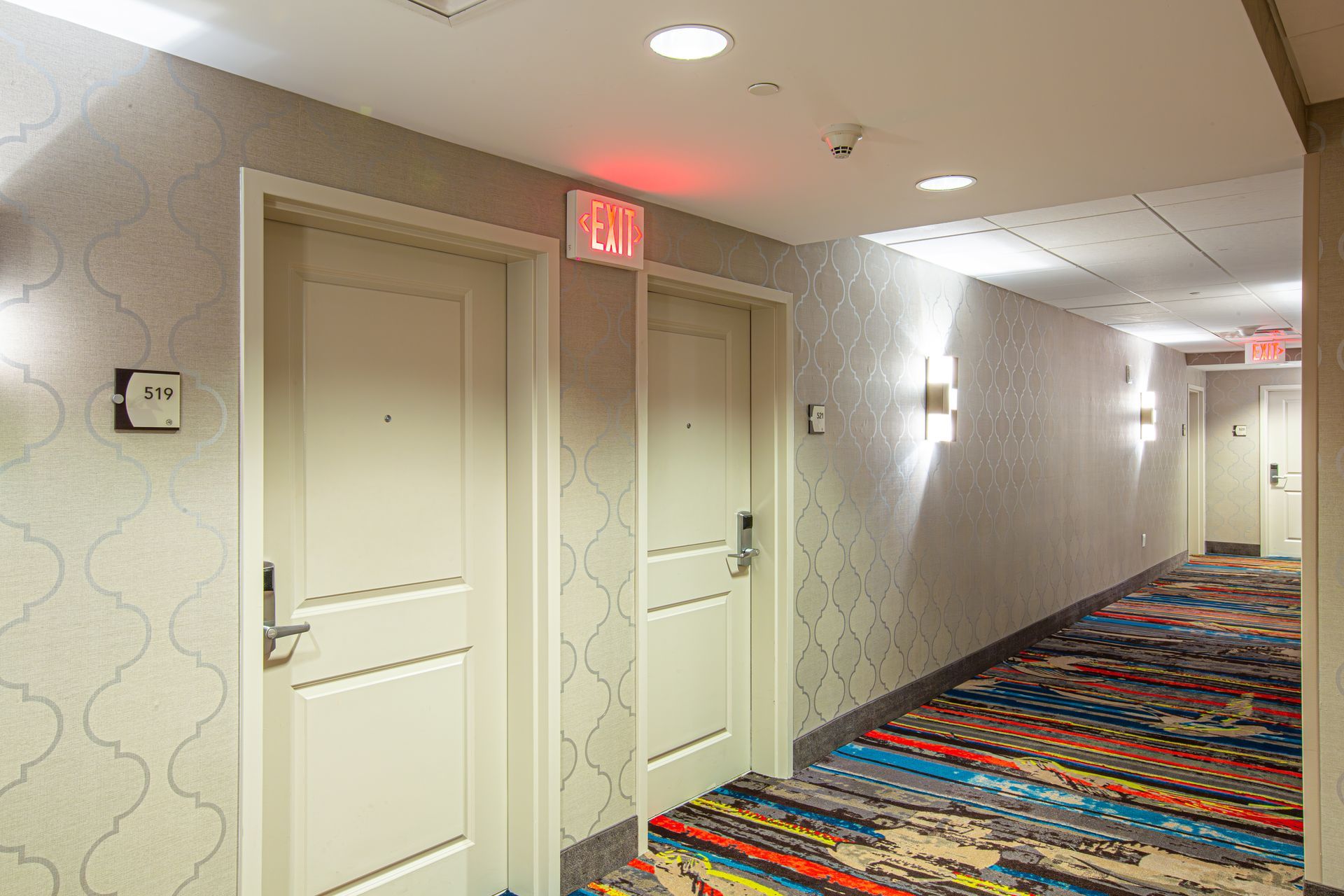 Hotel hallway with doors, exit sign, patterned carpet, and wall sconces.