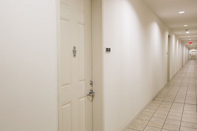 White hallway with doors, overhead lights, and a tiled floor. An exit sign is visible.