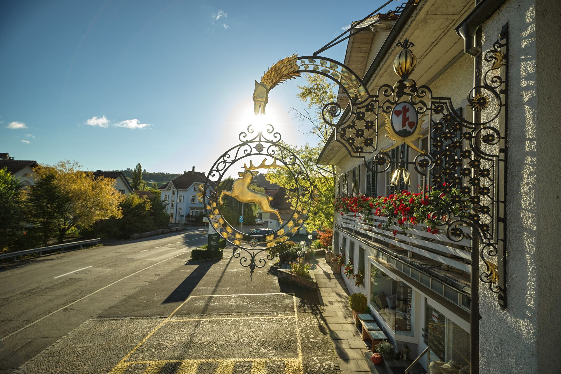 Sonnendurchflutete Straße mit dekorativem Torbogen und blumengeschmücktem Balkon neben einem malerischen Gebäude.