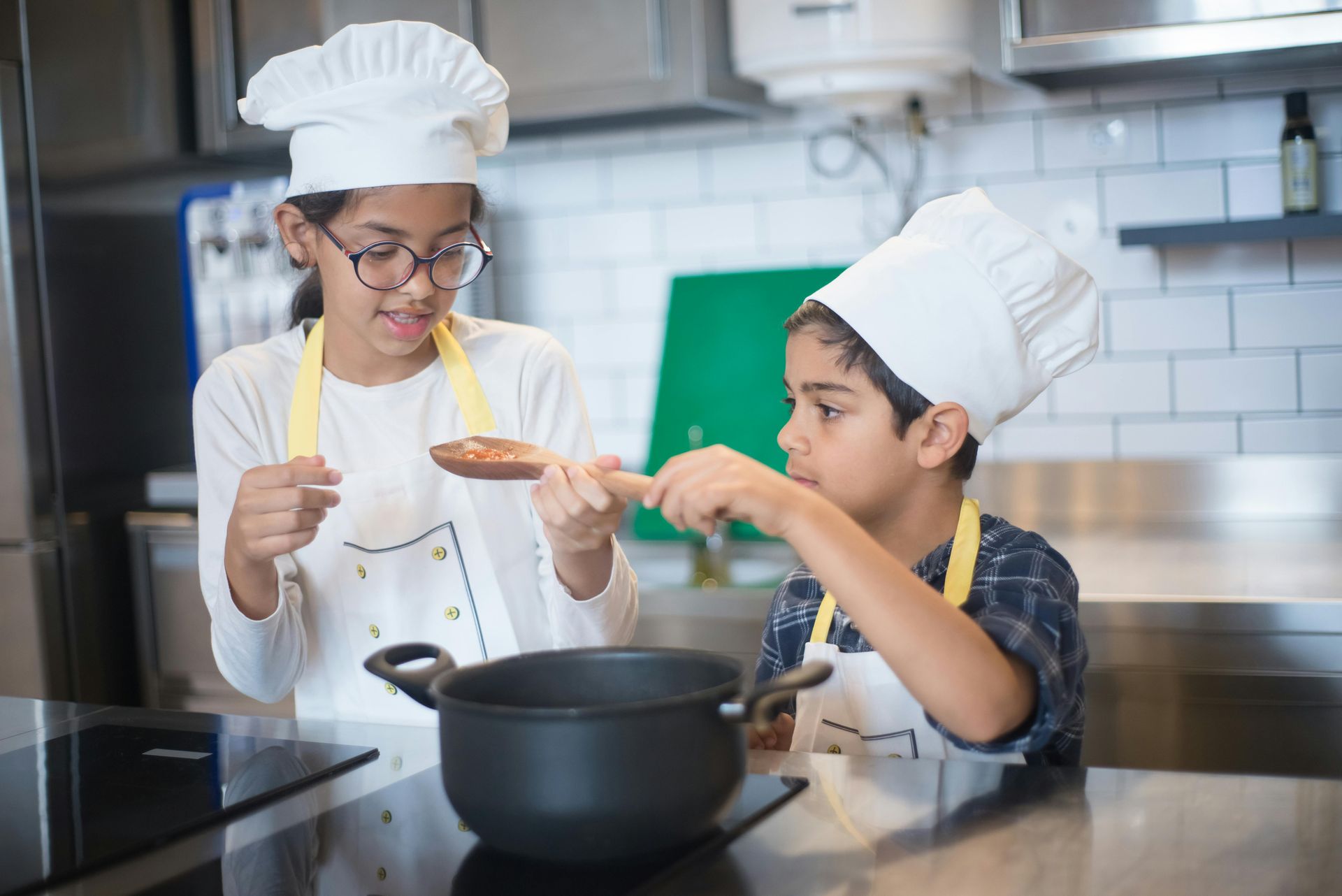 Zwei Kinder mit Kochmützen kochen gemeinsam in einer Küche, rühren in einem Topf und probieren das Essen.