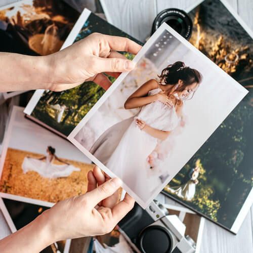 Hands holding a wedding photo over a spread of printed photos and a camera on a dark surface