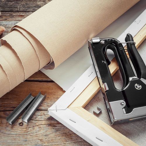 Staple gun beside wooden frame, nails, and rolled canvas on a worktable