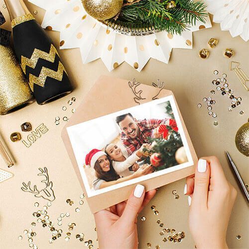 Hands holding a holiday photo card on a festive gold tabletop with ornaments and confetti