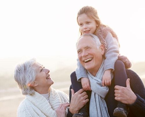 Smiling family outdoors at sunset, with a child on a man's shoulders and an older woman beside them