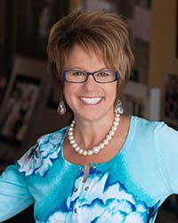Smiling woman in glasses and pearl necklace wearing a blue floral blouse indoors
