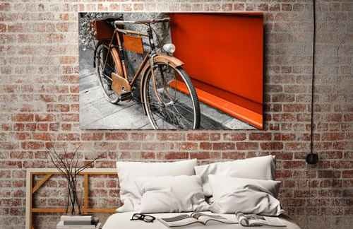 Framed bicycle photo above a bed against a brick wall with orange accent panel