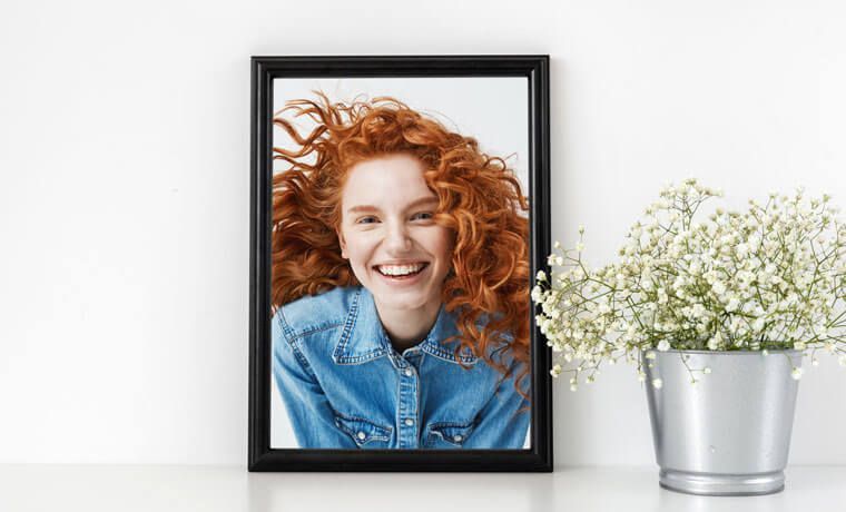 Framed portrait of a smiling red-haired person beside a vase of white flowers on a white surface