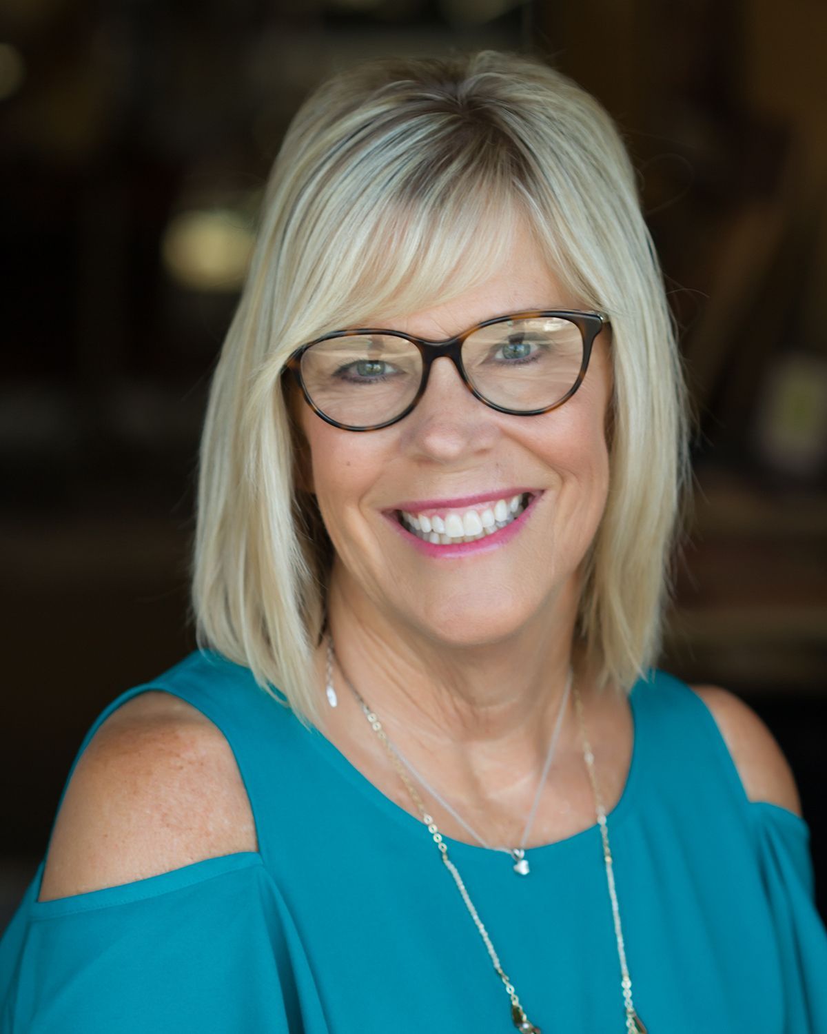 Smiling woman with blonde hair, glasses, and teal cold-shoulder top against a dark background