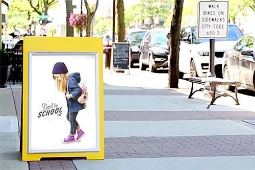 Yellow sidewalk sign advertising school with a child wearing a backpack, on a city street.