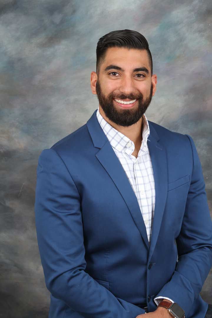 Smiling man in a blue suit and white checkered shirt against a gray studio backdrop