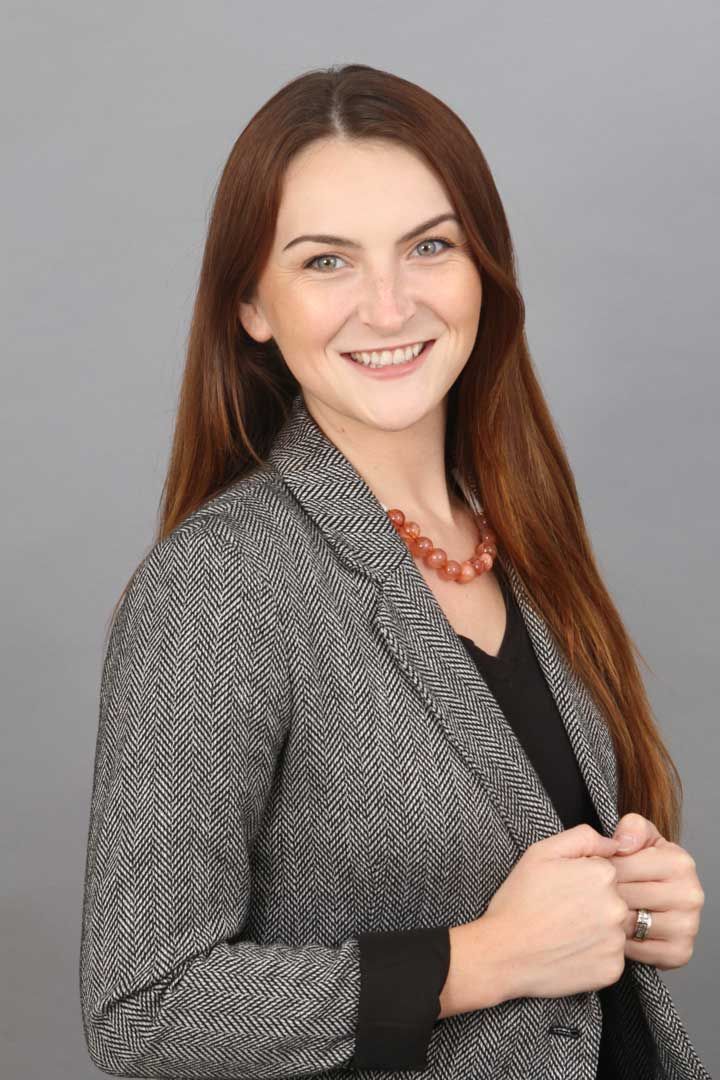 Smiling woman in a gray blazer and coral necklace against a gray backdrop