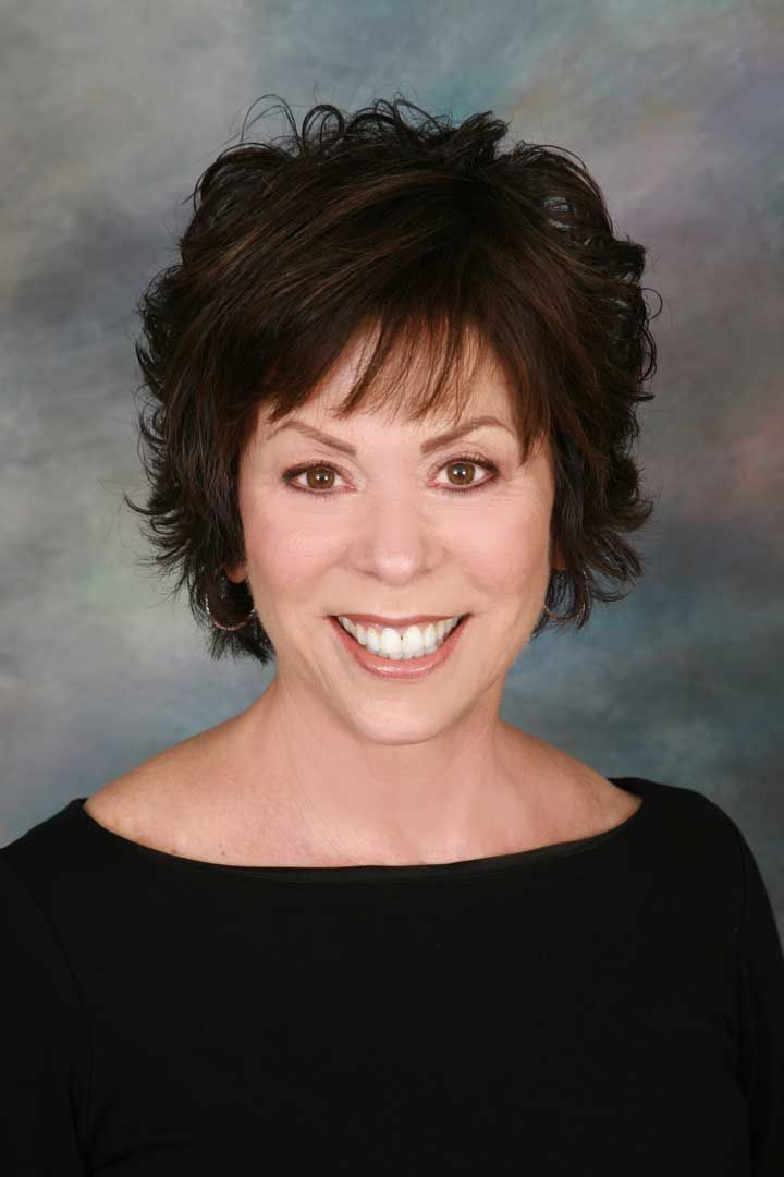 Smiling person with short dark curly hair in a black top against a gray studio background