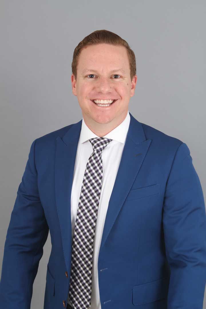 Man in a blue suit and checkered tie smiling against a plain gray background