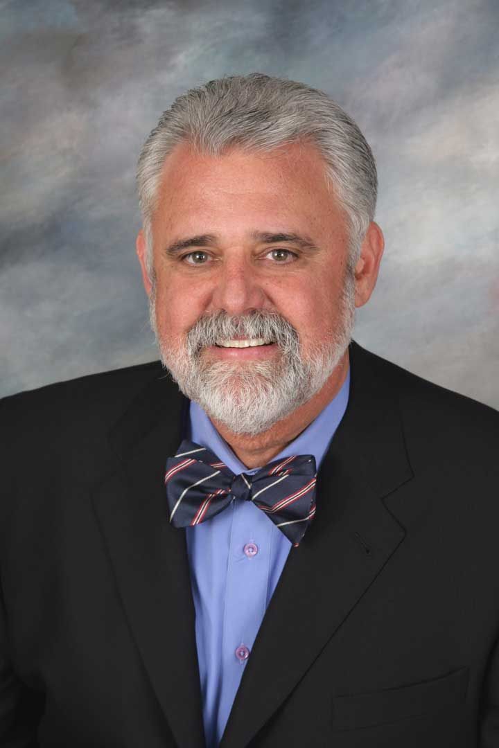 Portrait of a smiling man in a dark suit, blue shirt, and bow tie against a gray studio backdrop