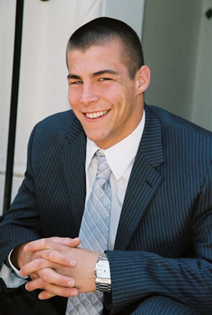 Smiling man in a dark pinstripe suit seated with clasped hands near a doorway