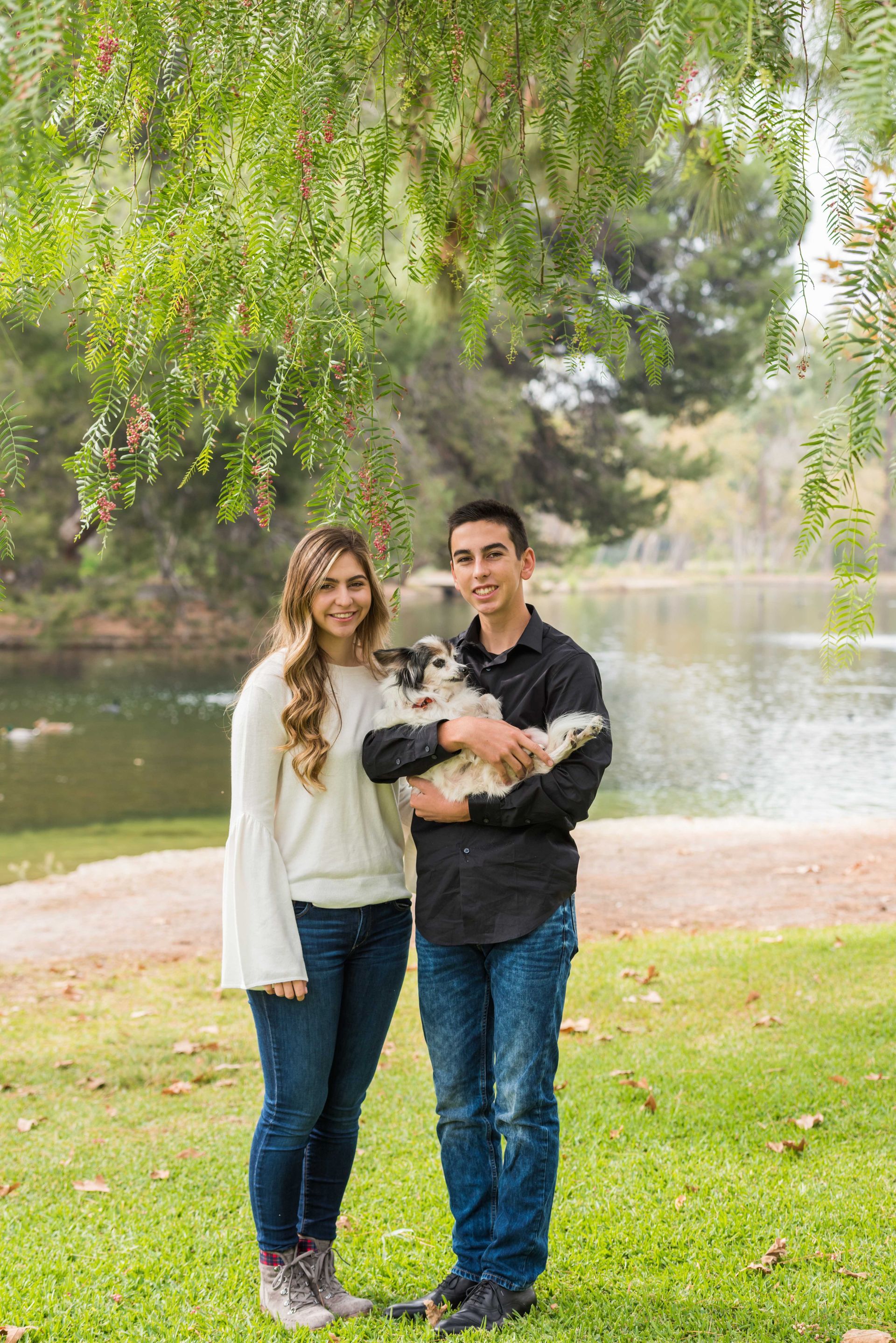 Two people holding a black-and-white dog beside a lake in a green park