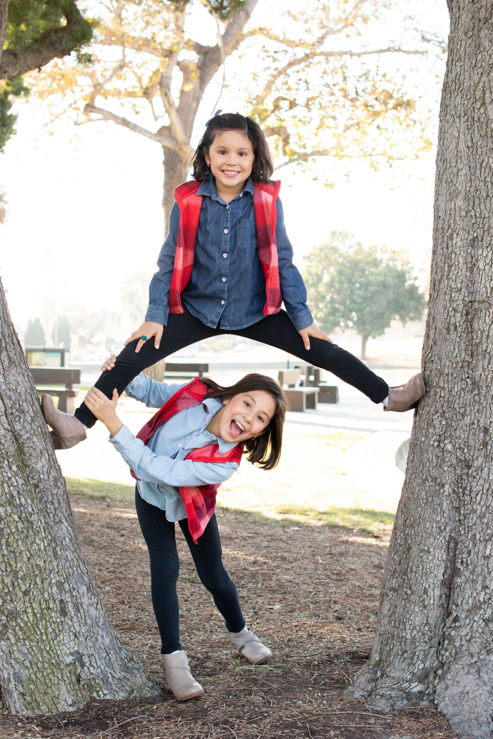 Two children posing playfully between two trees in a park, one standing on the other’s shoulders.