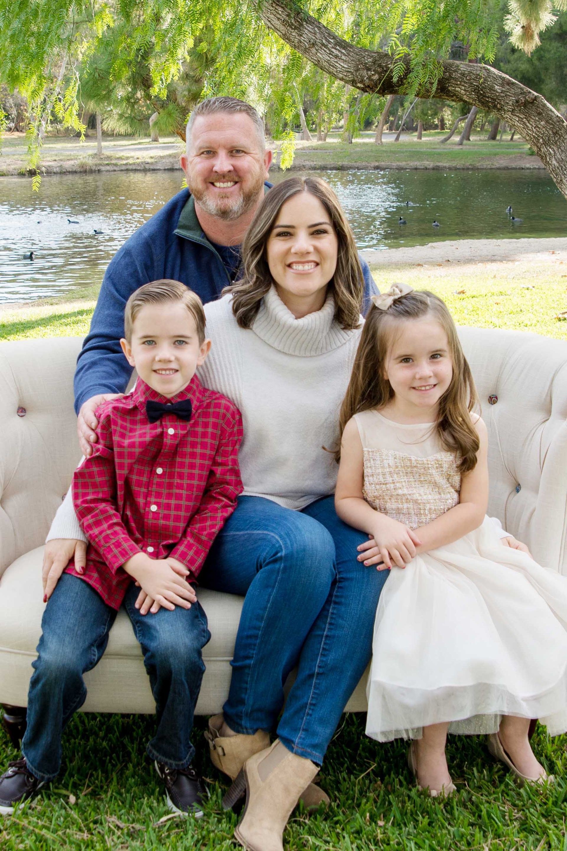 Family of four sitting on a white couch outdoors by a pond, smiling at the camera.