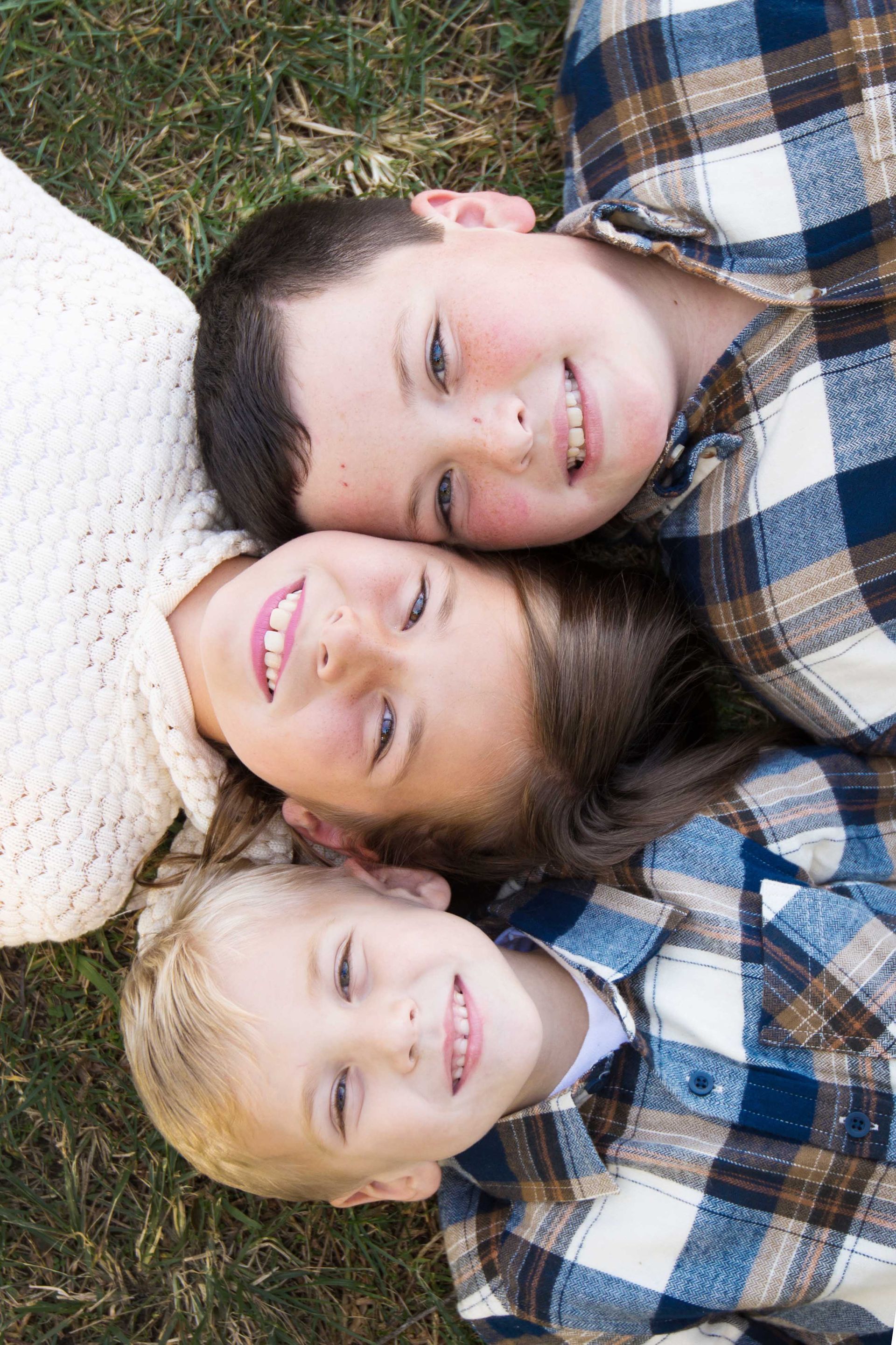 Three smiling children lying on grass, heads together, wearing plaid shirts and a cream sweater
