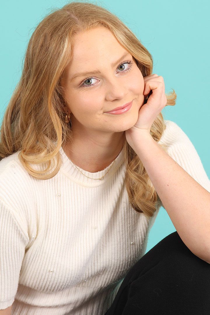 Smiling person with curly blonde hair resting chin on hand against a teal background