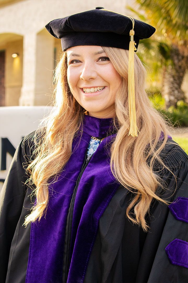 Smiling graduate in black cap and gown with purple stole, outdoors in sunlight