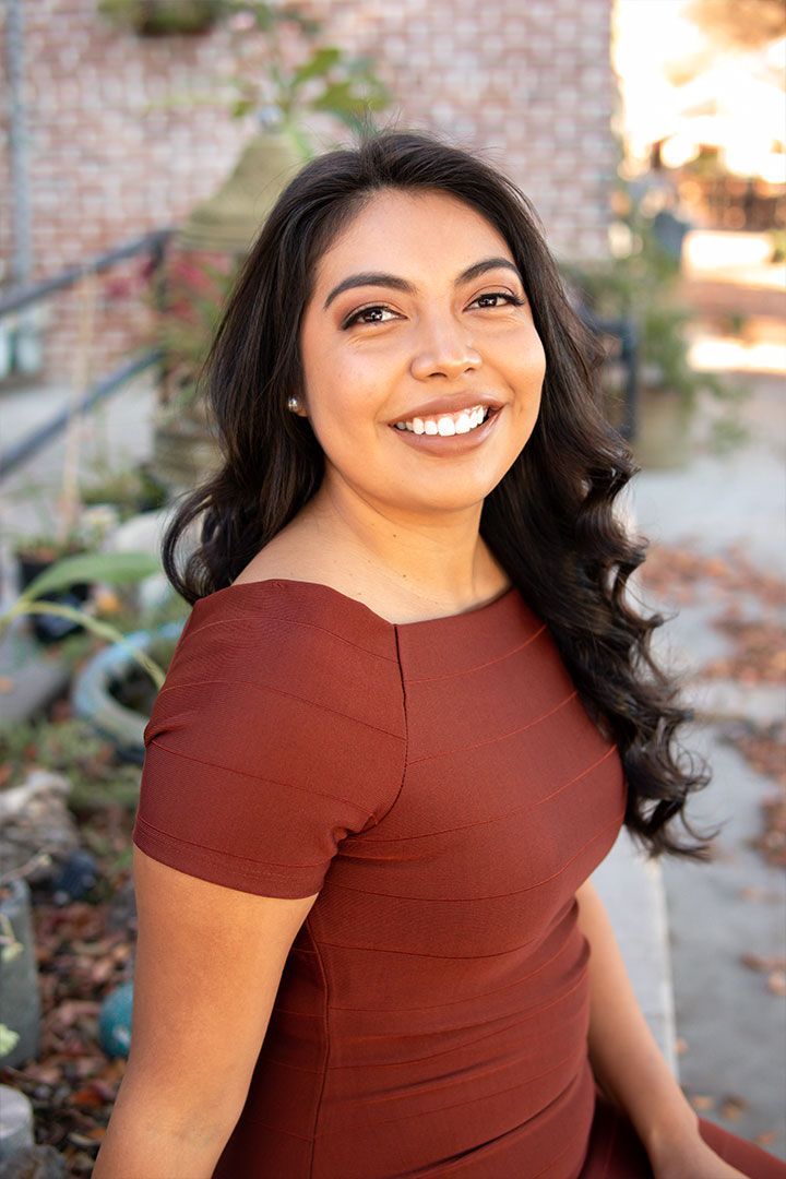 Smiling woman in a rust-colored dress standing outdoors on a brick path.