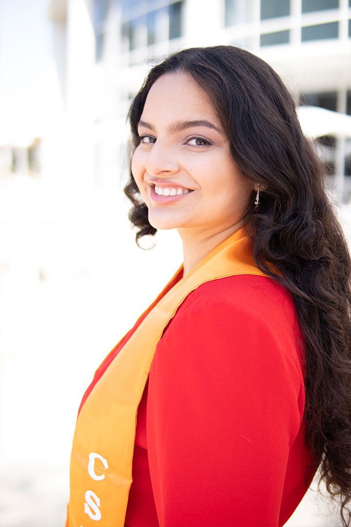 Smiling woman in a red top and orange scarf, standing outdoors against a bright building background
