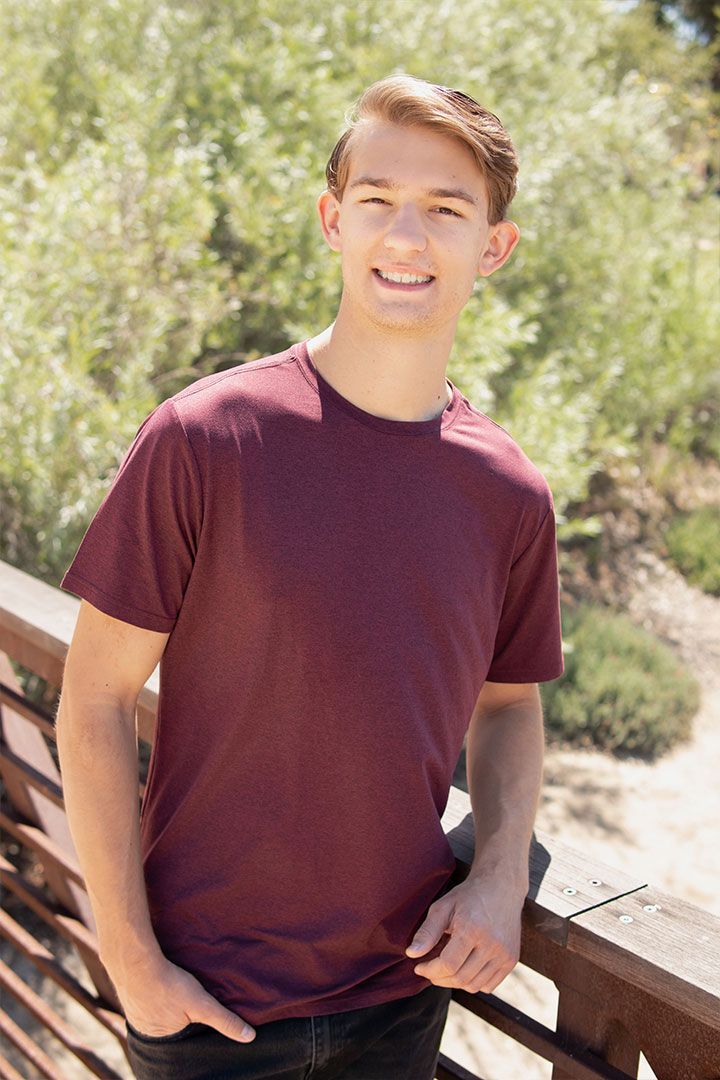 Smiling person in a maroon T-shirt leaning on a wooden railing outdoors in sunlight