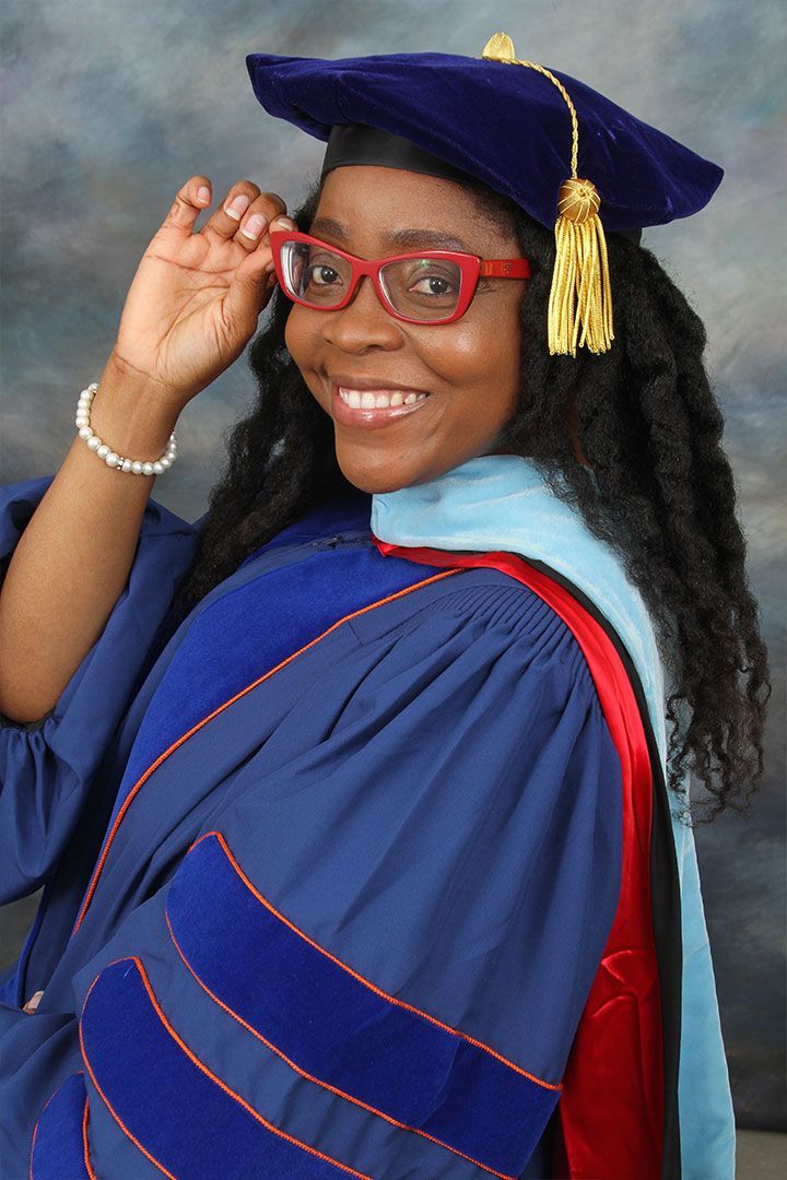 Graduate in blue cap and gown, smiling and adjusting red glasses against a studio backdrop.