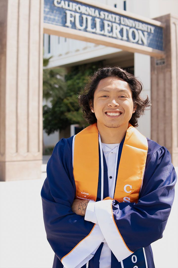 Graduate in blue gown and orange stole standing in front of a California State University, Fullerton sign.