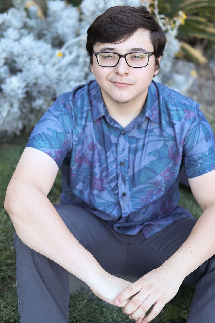 Person in a blue patterned shirt and glasses sitting outdoors beside a bush, looking at the camera.