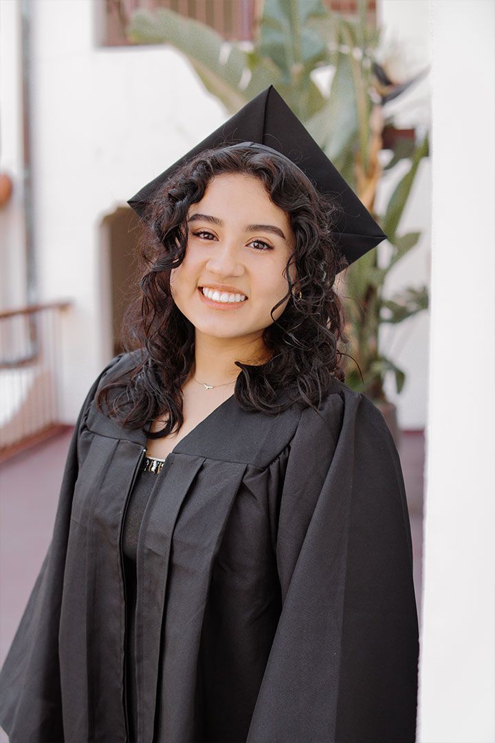 Person in a black graduation cap and gown smiling outdoors.