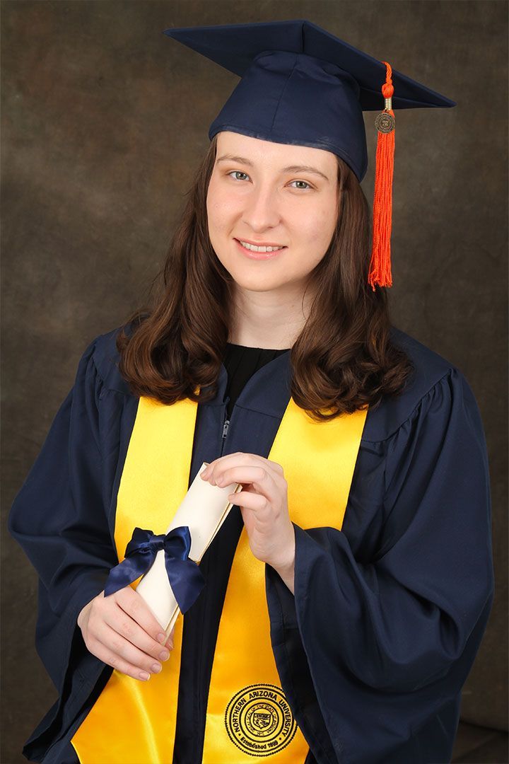 Graduate in navy cap and gown holding diploma, wearing gold stole and smiling against a dark backdrop