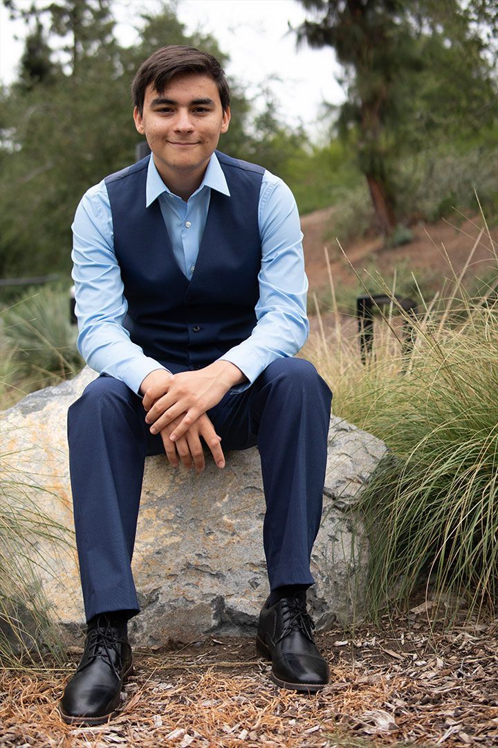Person in a blue shirt and vest sits on a rock outdoors in a natural setting.