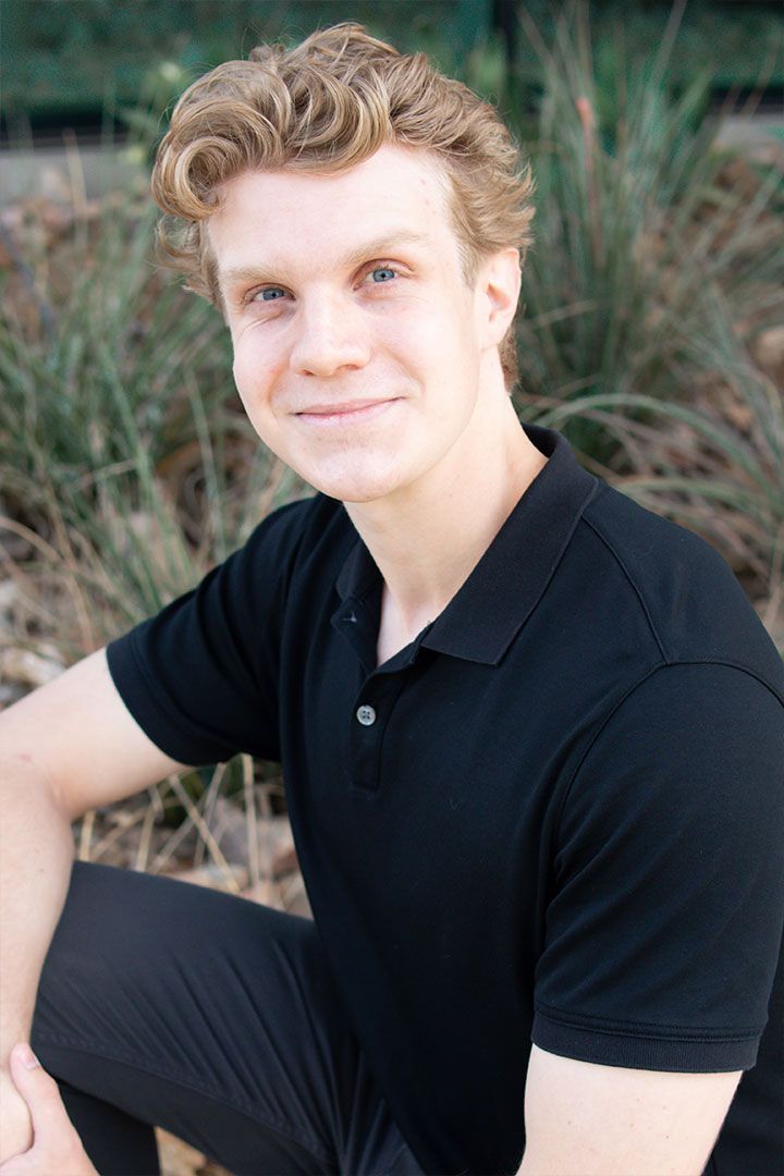 Smiling person in a black polo shirt sitting outdoors with greenery in the background