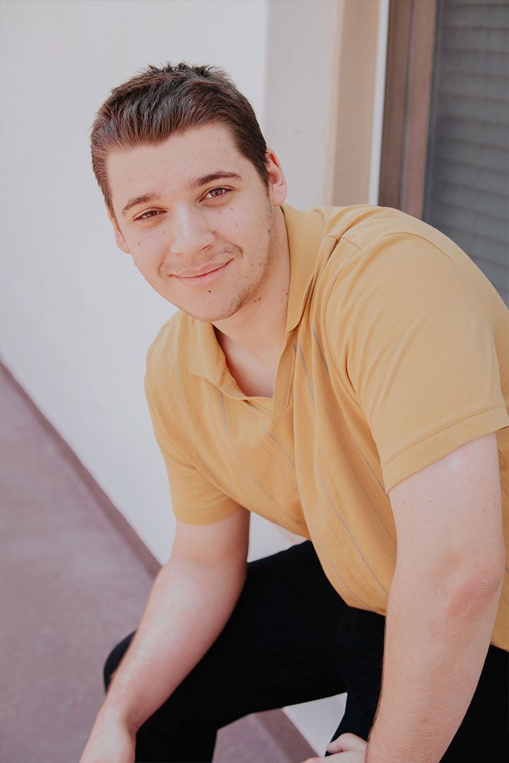 Smiling person in a yellow shirt sitting outdoors against a light wall.
