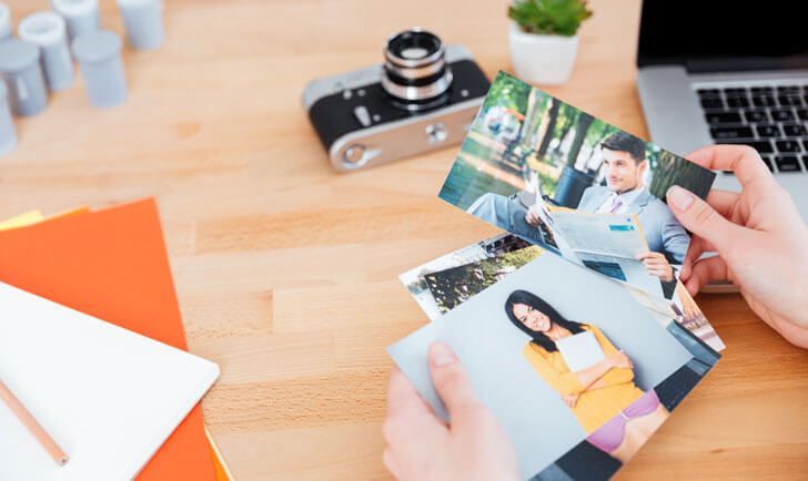 Hands holding printed photos over a desk with camera, notebook, and laptop keyboard in the background