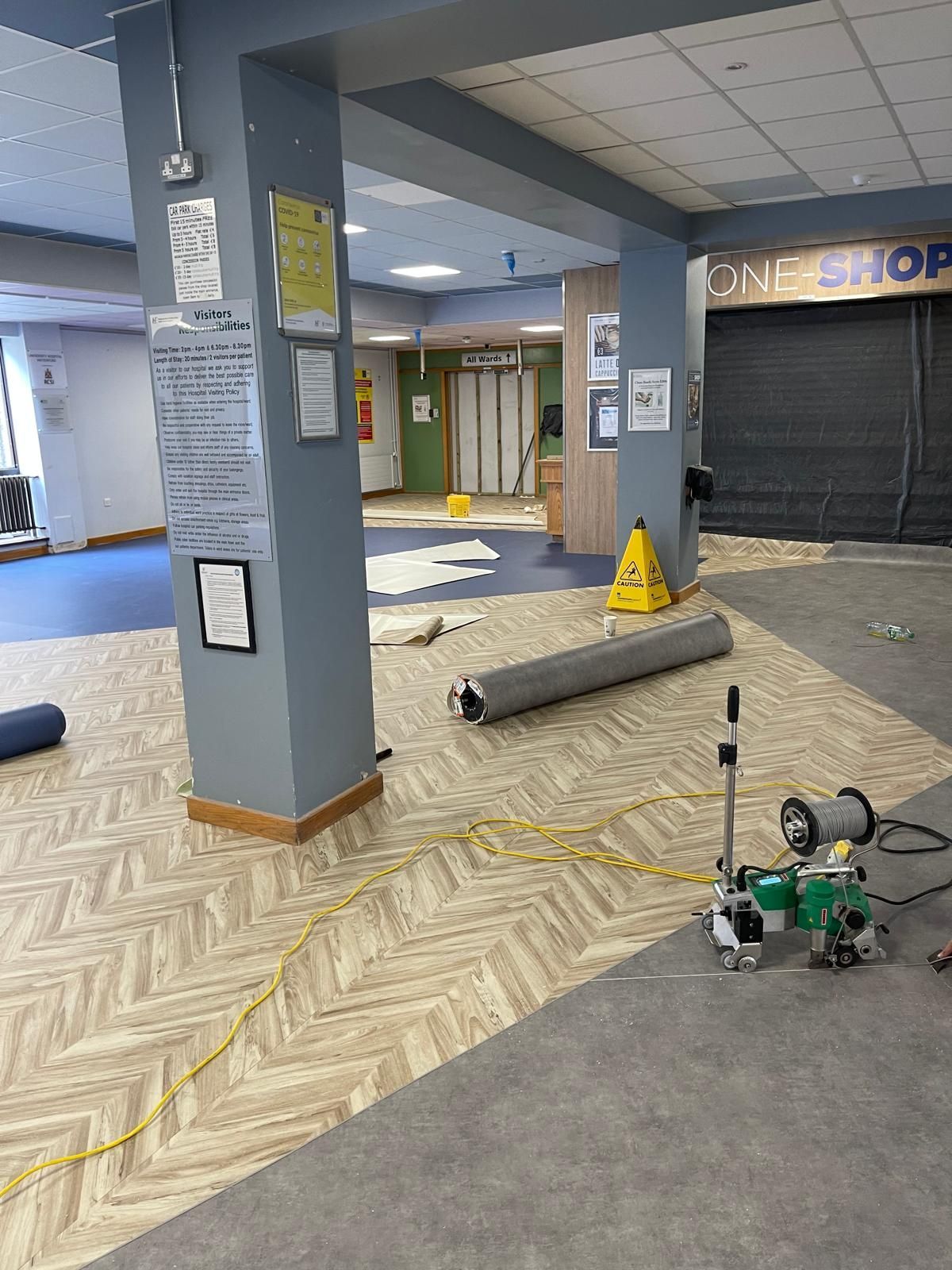 Interior space during renovation: new flooring being installed. Rolling carpet, machinery, elevator, caution signs visible.