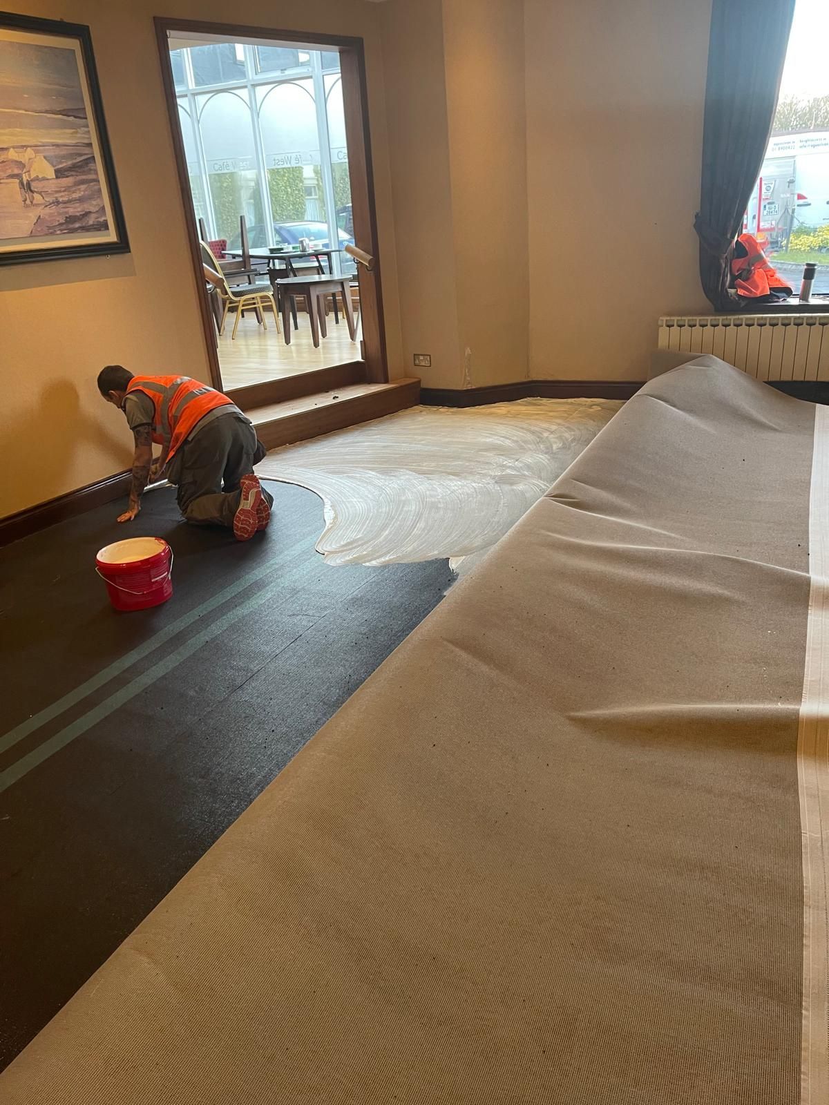A person in an orange vest applies adhesive on a floor, installing carpet in a room with a door to a sunroom.