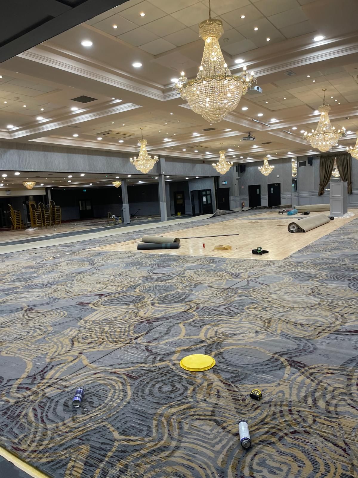 Large ballroom with ornate chandeliers and patterned carpet, under construction with rolls of carpet and equipment.