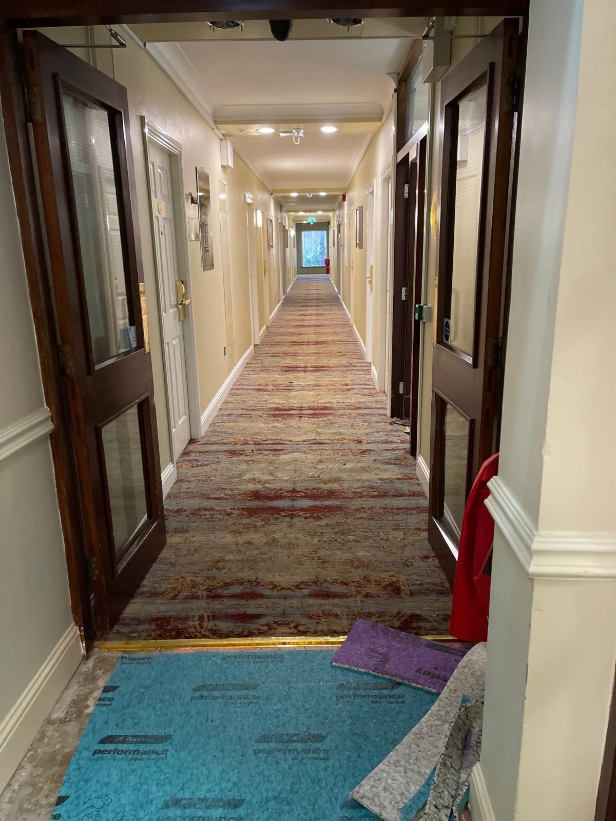 Hotel hallway with doors and patterned carpet. Teal mat and red coat near foreground doors.