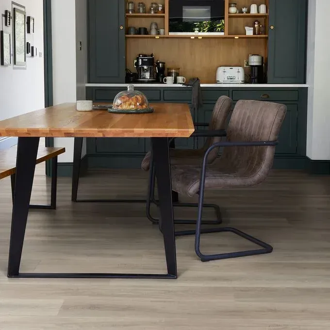 A wooden dining table with a black metal base and two modern brown chairs, set against a dark kitchen background.