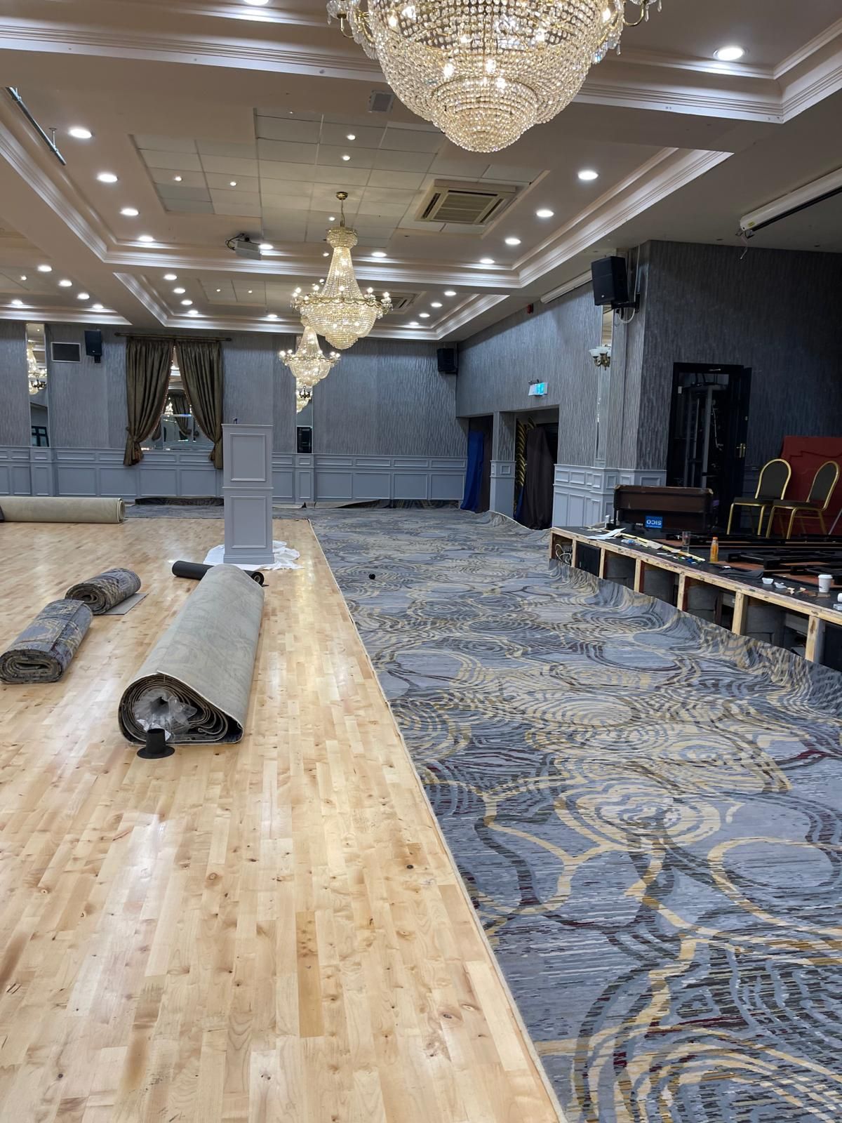A large room with wooden floor and carpet being installed. Chandelier lighting.
