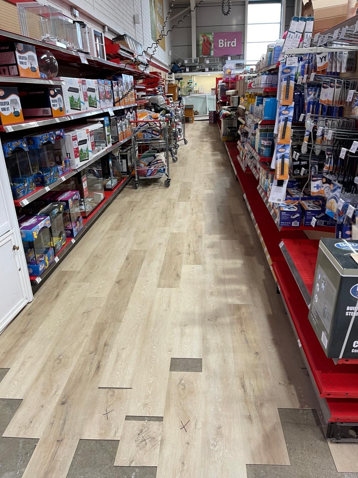 Interior view of a store aisle with shelves filled with products, light wood-look flooring, and overhead lighting.