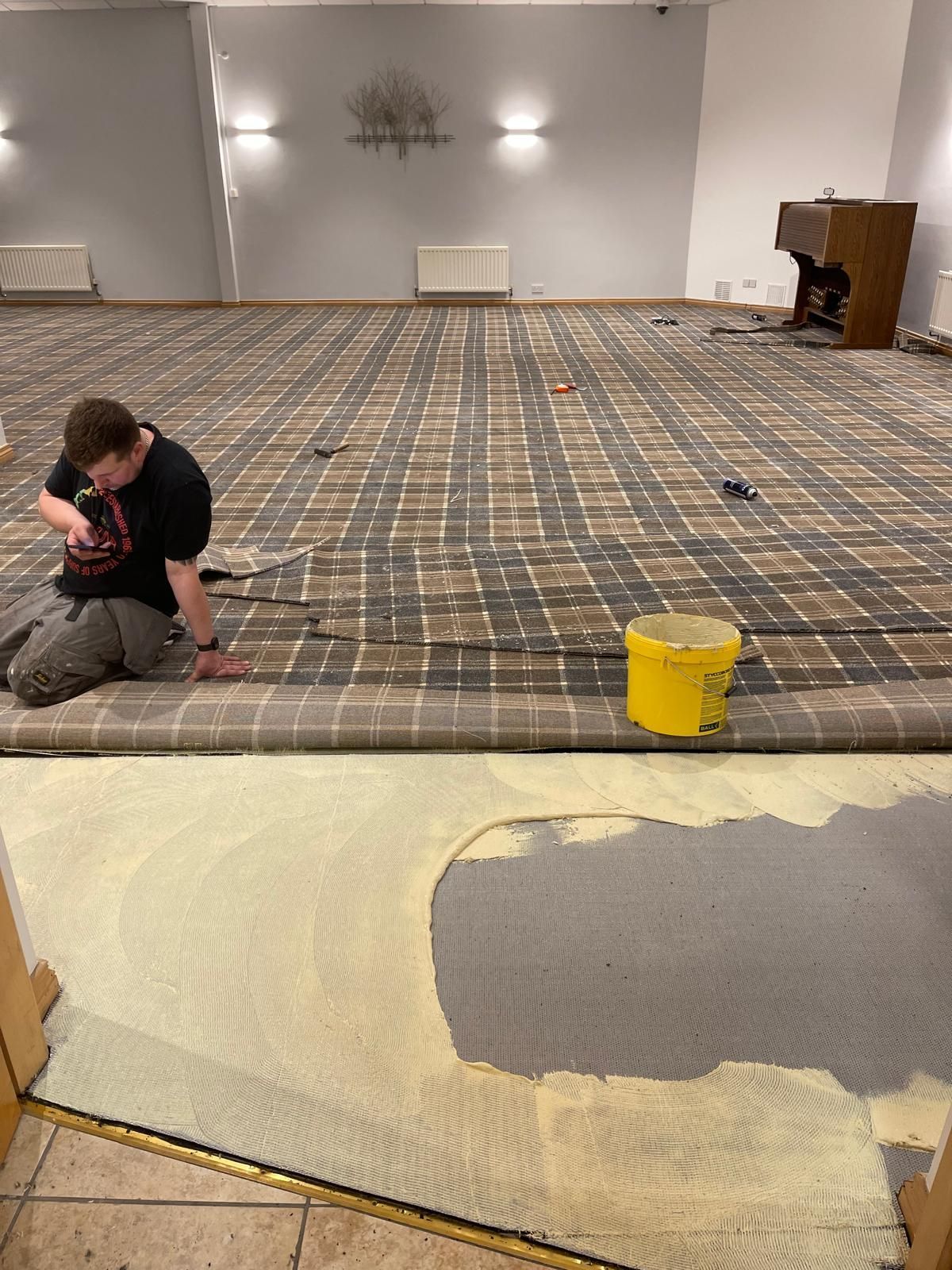 Person installing carpet in a room with tan and brown plaid flooring, a yellow bucket, and a partially carpeted floor.