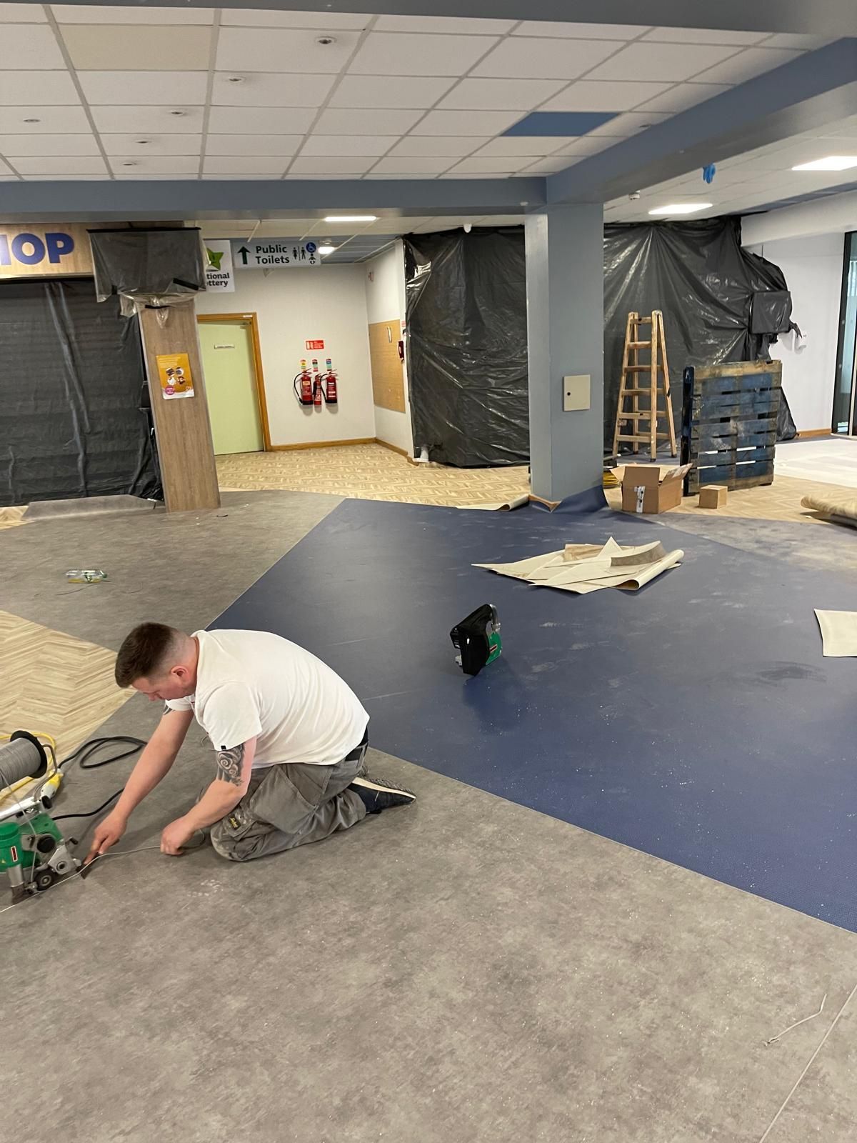 Man installing flooring in a commercial space, with blue and grey flooring, construction materials visible.